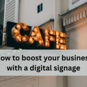 A vintage style cafe sign with glowing round bulbs hangs on the exterior of a light colored building with shutters