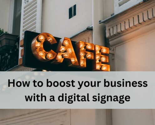 A vintage style cafe sign with glowing round bulbs hangs on the exterior of a light colored building with shutters