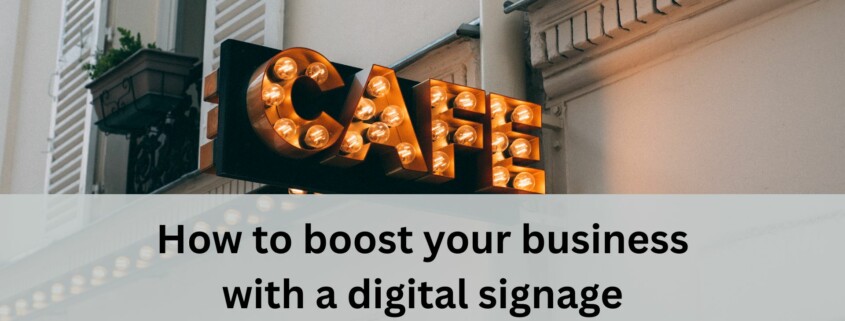 A vintage style cafe sign with glowing round bulbs hangs on the exterior of a light colored building with shutters