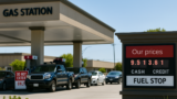 Gas station with cars in line and a roadside sign showing separate cash and credit fuel prices.