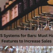Rows of hanging wine glasses at a bar with bottles on shelves above, along with the text “POS Systems for Bars: Must Have Features to Increase Sales”