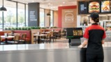 Restaurant employee using a touchscreen POS system in a modern fast-food restaurant with dining tables, booths, digital menu boards, and counter service.