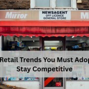 Front view of a small retail store with a red awning, newspaper signage, and various products visible through the windows