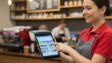 A smiling server in a red shirt and grey apron holds a free point of sale device, taking an order in a coffee shop.